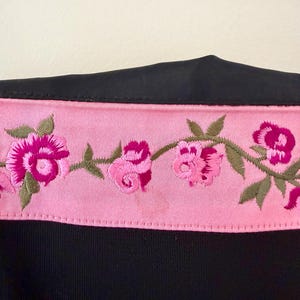 Puede incluir: Una prenda negra con un ribete bordado floral rosa. El ribete presenta rosas rosas y hojas verdes. La tela de la prenda parece ser un material liso, posiblemente satinado.