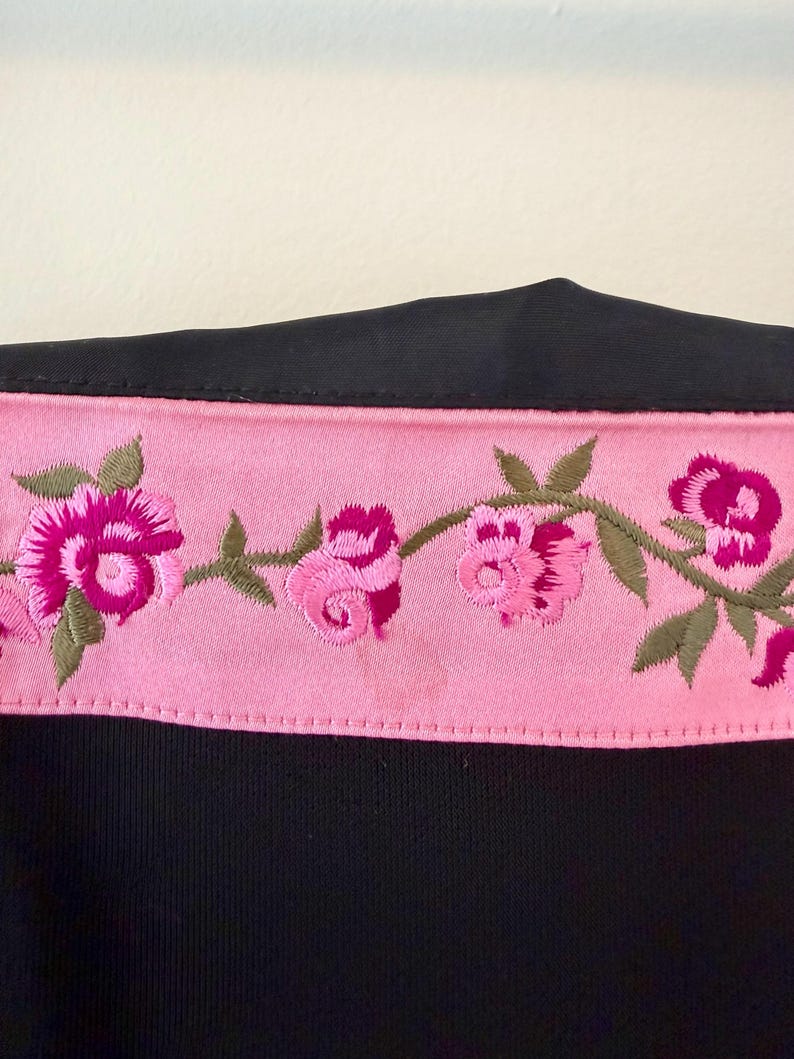 Puede incluir: Una prenda negra con un ribete bordado floral rosa. El ribete presenta rosas rosas y hojas verdes. La tela de la prenda parece ser un material liso, posiblemente satinado.
