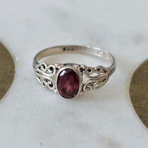 May include: A silver ring with an oval, deep red gemstone. The ring features an ornate, filigree design on the band, with decorative swirls on either side of the gemstone. The ring is set on a white and gold surface.