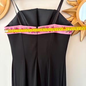 Puede incluir: Vestido de noche negro con tirantes finos. El vestido presenta un detalle de cinta rosa con bordado floral a lo largo del borde superior. La cinta mide aproximadamente 25 cm de ancho. El vestido est&aacute; colgado en una percha de madera.