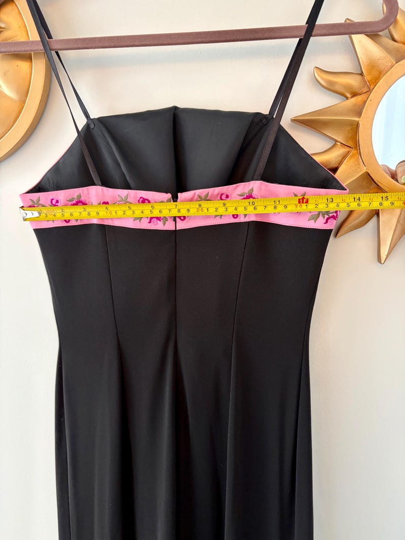 Puede incluir: Vestido de noche negro con tirantes finos. El vestido presenta un detalle de cinta rosa con bordado floral a lo largo del borde superior. La cinta mide aproximadamente 25 cm de ancho. El vestido est&aacute; colgado en una percha de madera.