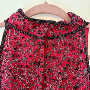K&ouml;nnte beinhalten: Ein &auml;rmelloses rotes Oberteil mit aufwendiger schwarzer Perlenarbeit und silbernen Akzenten. Das Design zeigt ein Blumenmuster mit Bl&auml;ttern und Beeren aus Perlen. Der Ausschnitt und die Kanten sind mit schwarzen Perlen verziert.