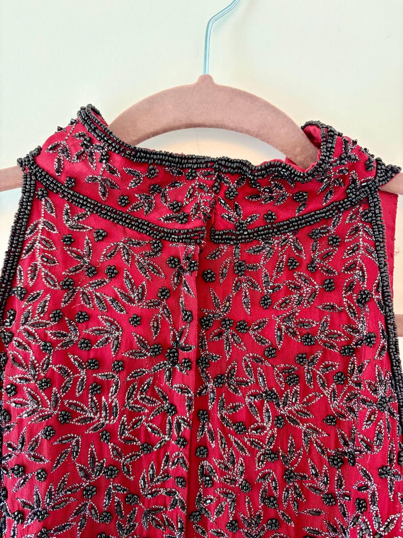K&ouml;nnte beinhalten: Ein &auml;rmelloses rotes Oberteil mit aufwendiger schwarzer Perlenarbeit und silbernen Akzenten. Das Design zeigt ein Blumenmuster mit Bl&auml;ttern und Beeren aus Perlen. Der Ausschnitt und die Kanten sind mit schwarzen Perlen verziert.