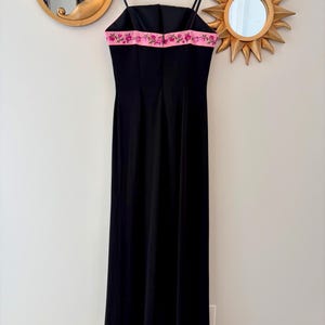Puede incluir: Un vestido de noche negro con tirantes finos y falda larga. El vestido presenta una banda rosa con bordados florales en el escote y el dobladillo. Cuelga de una percha de madera contra una pared neutra.