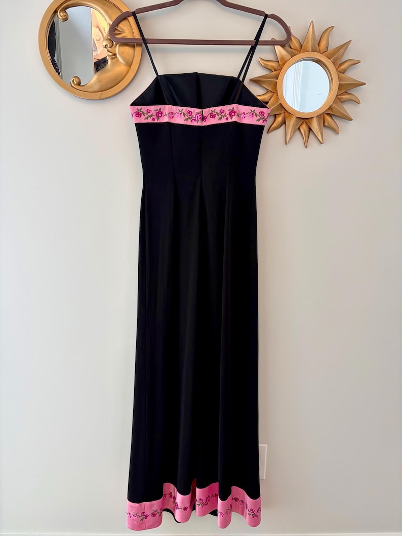 Puede incluir: Un vestido de noche negro con tirantes finos y falda larga. El vestido presenta una banda rosa con bordados florales en el escote y el dobladillo. Cuelga de una percha de madera contra una pared neutra.