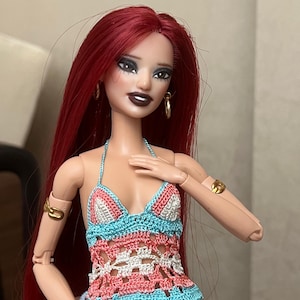 May include: A doll with long red hair wearing a handmade crochet top. The top features a halter neck and a pattern of white, coral, and turquoise. The doll also has gold hoop earrings and a gold bracelet.