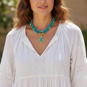 May include: A double-strand necklace featuring round turquoise beads and gold-toned metal spacers. The necklace includes a central teardrop-shaped turquoise pendant set in a gold-toned frame, resting against a white long-sleeved blouse with lace.