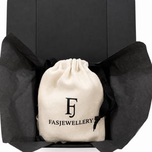 May include: A cream-colored fabric drawstring pouch with black text reading FASJEWELLERY and a stylized FJ logo, nestled inside an open black gift box lined with black tissue paper. The pouch features a black drawstring cord at the top.