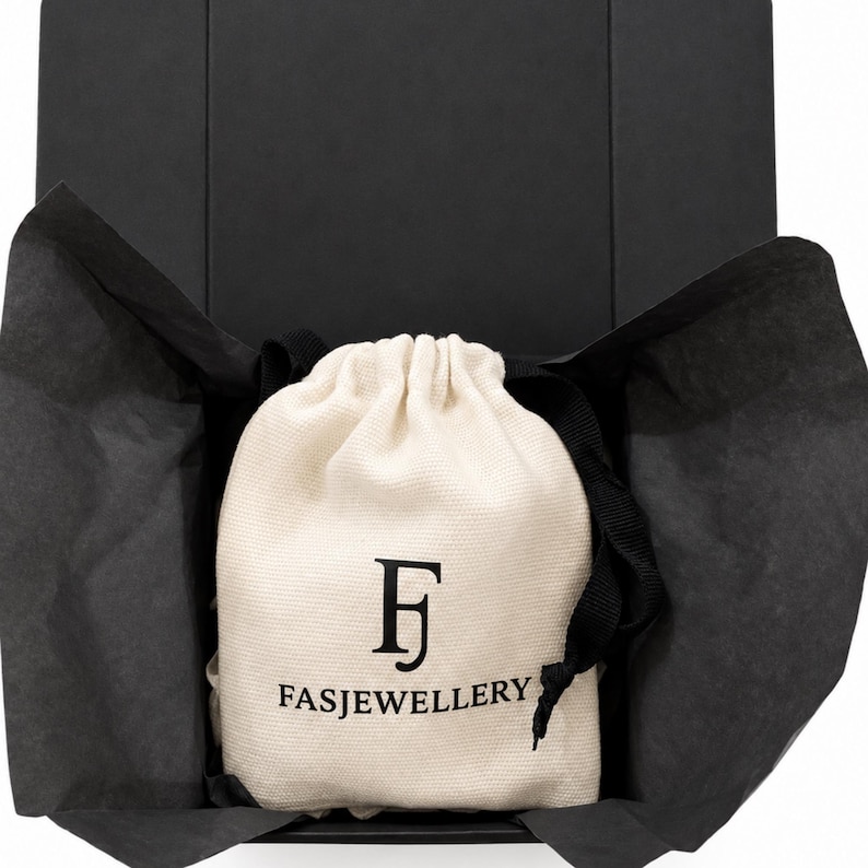 May include: A cream-colored fabric drawstring pouch with black text reading FASJEWELLERY and a stylized FJ logo, nestled inside an open black gift box lined with black tissue paper. The pouch features a black drawstring cord at the top.