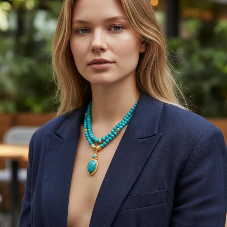 May include: A double-strand turquoise beaded necklace featuring round beads and a central teardrop-shaped turquoise pendant set in a gold-toned metal frame with decorative gold-toned spacers, worn over a dark navy blue blazer with a notched lapel.