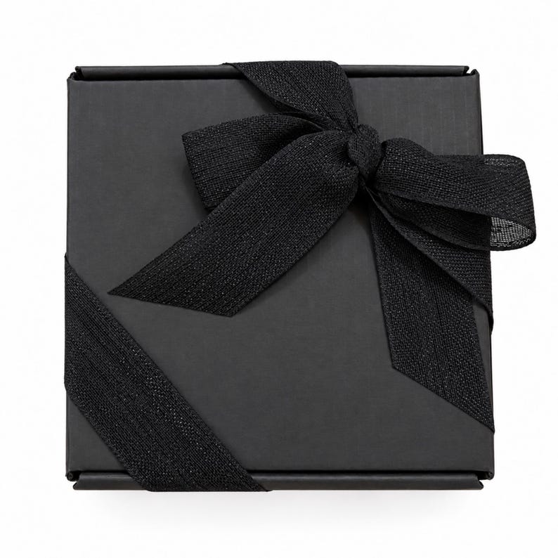 May include: A square black gift box featuring a matte finish, wrapped with a textured black ribbon tied into a decorative bow on the top surface. The ribbon extends diagonally across the box, creating a clean and uniform appearance.