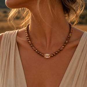 Pode incluir: Um colar com uma série de contas facetadas de cor bronze dispostas num padrão circular. O centro do colar apresenta um único pendente de cristal retangular de cor champanhe claro, criando um contraste com as contas escuras.