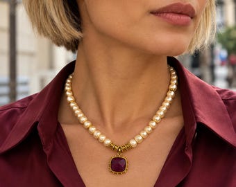 May include: A necklace featuring a strand of cream-colored pearls interspersed with small gold-toned metal beads. The necklace includes a central square-shaped pendant with a deep magenta faceted stone set in a gold-toned frame.
