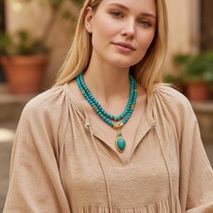 May include: A double-strand necklace featuring round turquoise-colored beads and gold-toned metal accents. The necklace includes a central teardrop-shaped turquoise-colored pendant framed in gold-toned metal, resting against a beige textured blouse.