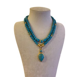 May include: A double-strand necklace featuring round turquoise-colored beads interspersed with gold-toned metal spacers. The necklace includes a central pendant with a small round bead above a larger teardrop-shaped stone in a gold-toned setting.