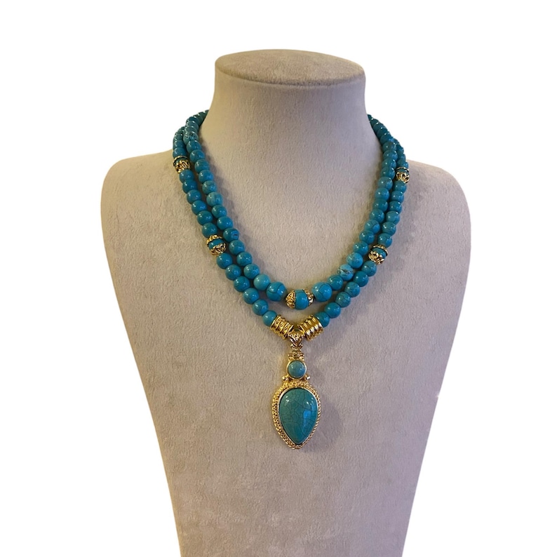 May include: A double-strand necklace featuring round turquoise-colored beads interspersed with gold-toned metal spacers. The necklace includes a central pendant with a small round bead above a larger teardrop-shaped stone in a gold-toned setting.