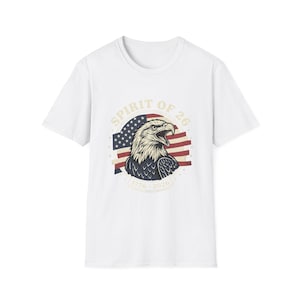 May include: White t-shirt featuring a graphic of an eagle with an American flag design. The text "SPIRIT OF 26" is arched above the eagle, with the years "1776 - 2026" below. The eagle is in shades of brown, white, and blue.