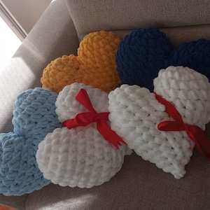 Könnte beinhalten: Eine Sammlung herzförmiger Kissen in verschiedenen Farben, darunter Gelb, Blau und Weiß. Zwei weiße Kissen sind mit roten Bändern gebunden. Die Kissen bestehen aus einem weichen, strukturierten Material und sind auf einem neutralfarbenen Sofa angeordnet.