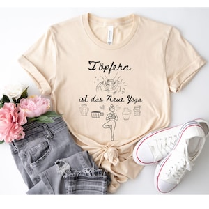 Könnte beinhalten: Beiges T-Shirt mit dem schwarzen Schriftzug "Töpfern ist das Neue Yoga". Das Shirt zeigt Illustrationen von Töpferwerkzeugen und einer Yoga-Pose. Das Shirt wird mit grauen Jeans und weißen Turnschuhen kombiniert.