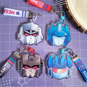Könnte beinhalten: Vier Acryl-Schlüsselanhänger mit stilisierten Charakterköpfen aus der Transformers-Reihe, darunter Optimus Prime und Megatron. Jeder Anhänger ist an einer Stoffschlaufe mit einem Metallkarabiner befestigt und mit den Namen der Figuren bedruckt.