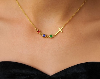 Puede incluir: Un collar dorado con un colgante de cruz y cuatro piedras preciosas de colores: rojo, azul y verde. El collar se lleva alrededor del cuello, con las piedras dispuestas en una línea curva.