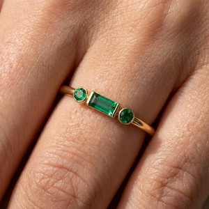 May include: A gold ring featuring three green emerald stones. The ring has a rectangular emerald in the center, flanked by two round emeralds. The band is a thin, polished gold.