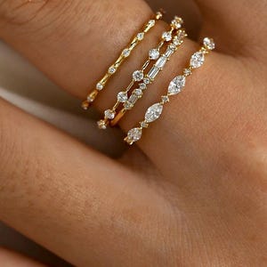 May include: Four gold rings with diamond detailing are shown on a finger. The rings feature different diamond cuts, including round, baguette, and marquise shapes. The rings are set in gold bands, creating a delicate and elegant appearance.