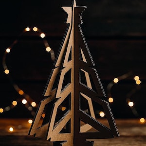 Könnte beinhalten: Ein dekorativer Weihnachtsbaum aus braunem Karton mit einem Stern an der Spitze, vor einem dunklen Holzgrund. Der Baum hat ein ausgeschnittenes Design und wird von Lichterketten beleuchtet. Der Baum ist aus Pappe.