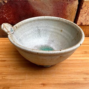 May include: A round, hand-thrown ceramic bowl with a speckled off-white glaze and a prominent, textured handle on one side. The interior base features a vibrant, circular patch of turquoise-green glaze, contrasting with the neutral exterior.