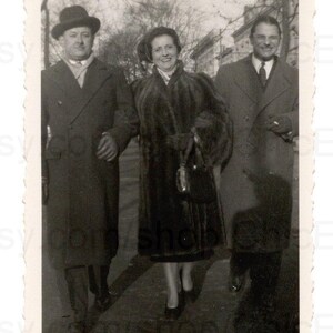 Op de afbeelding: Vintage zwart-witfoto van drie mensen die buiten lopen. Een vrouw in een bontjas wordt geflankeerd door twee mannen in overjassen en hoeden. De afbeelding heeft een klassieke, tijdloze uitstraling.