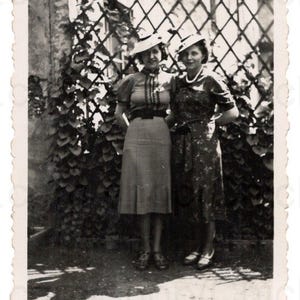 Op de afbeelding: Vintage zwart-witfoto met twee vrouwen die buiten poseren. Ze dragen hoeden en jurken, staand voor een trellis bedekt met bladeren. De foto heeft een geschulpte rand, wat bijdraagt aan de antieke uitstraling.