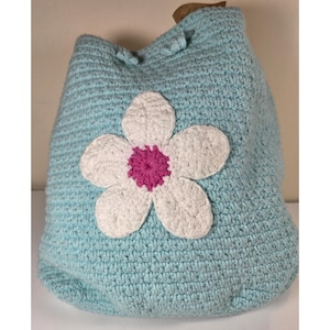 May include: A light blue crochet tote bag with a large white and pink flower applique. The bag has a drawstring closure and a brown handle. The bag is handmade and has a textured appearance.