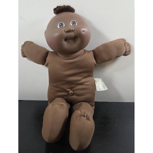 May include: A brown Cabbage Patch Kids doll with a dark complexion. The doll has a tuft of brown hair and open arms. The doll is wearing a brown onesie and has stitched features. A tag is attached to the doll's side.