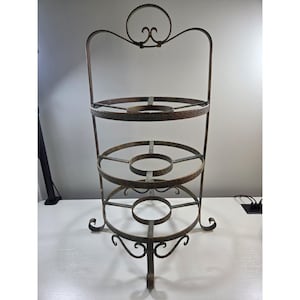 May include: A three-tiered metal stand with a dark, weathered bronze finish. The structure features circular rings on each level, supported by curved metal legs and a tall, arched handle at the top with a decorative circular loop detail.