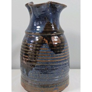 May include: A handcrafted ceramic pitcher with a dark blue and brown glaze. The pitcher has a ruffled rim and a textured, ribbed body. The colors blend together, creating a unique, rustic appearance. This decorative piece is suitable for home decor.