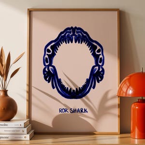 May include: A framed art print featuring a stylized, dark blue shark jaw on a light pink background. The text "ROR SHARK" is below the image. The print is displayed on a wooden cabinet with a red lamp and decorative vase.