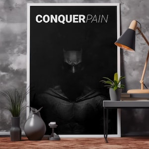 May include: A framed black and white poster with the text "CONQUER PAIN" above a shadowy image of a superhero. The poster is propped against a textured gray wall, with a small table and lamp to the right.