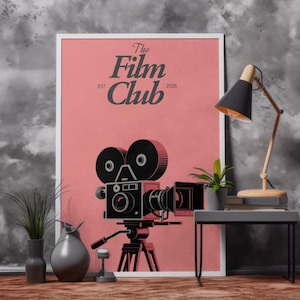 Peut inclure: Affiche encadrée avec un fond rose et une illustration d'une caméra de cinéma vintage. Le texte "The Film Club EST 2025" est affiché au-dessus de la caméra. Une lampe de bureau et une petite plante en pot sont sur une table d'appoint.