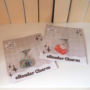 Könnte beinhalten: Zwei eReader-Anhänger auf einer Karte. Einer zeigt einen Stapel bunter Bücher, der andere eine rote Taschentuchbox. Beide Anhänger sind an einer Karte mit dem Text "eReader Charm" und dem Print & Paper Dreams-Logo befestigt.