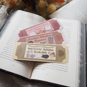 Könnte beinhalten: Drei dekorative Lesezeichen mit Herbstmotiven liegen auf einem offenen Buch. Die Lesezeichen tragen die Aufschriften "Cozy Autumn Library Ticket", "Sweater Weather Readers" und "Introverts' Autumn Bookclub". Getrocknete Blumen und Kürbiskerzen sind im Hintergrund.