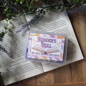 Könnte beinhalten: Ein rechteckiger, durchsichtiger Kunststoffbehälter mit Würfeln, versehen mit einem Etikett mit dem Text Reader's Roll The Cozy Reading Dice Game, einer Abbildung eines offenen Buches und violetten, orangefarbenen und weißen Wellen.