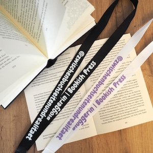 Könnte beinhalten: Zwei Schlüsselbänder mit dem Text "@zwischenbuchseitenundrealitaet | Bookish Press Bloggerin" werden auf einer Holzoberfläche neben einem offenen Buch gezeigt. Ein Schlüsselband ist schwarz mit weißem Text, das andere ist weiß mit lila Text.