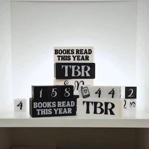 Könnte beinhalten: Eine Sammlung von schwarzen und weißen Holzblöcken mit verschiedenen Texten und Zahlen. Einige Blöcke zeigen "BOOKS READ THIS YEAR" und "TBR". Andere Blöcke zeigen Zahlen wie 1, 5, 8, 4 und 2. Die Blöcke sind auf einem weißen Regal angeordnet.