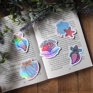 Könnte beinhalten: Fünf holografische Aufkleber mit ozeanischen Motiven, darunter Muscheln, ein Seestern und ein offenes Buch. Ein Aufkleber zeigt den Text "BOOK GIRL SUMMER" und ein weiterer den Text "STORIES IN A SEASHELL" auf einer Muschel.