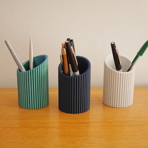 May include: Three cylindrical pen holders in teal, navy blue, and white, each with vertical ridged detailing. The teal holder contains a grey pen and a white pencil. The navy blue holder has several pens. The white holder has a black pen and a green pen.