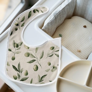 May include: A beige baby bib with a watercolor olive branch pattern, featuring green olives and leaves. The bib is on a white high chair tray, next to a divided plate and a cream-colored pillow.