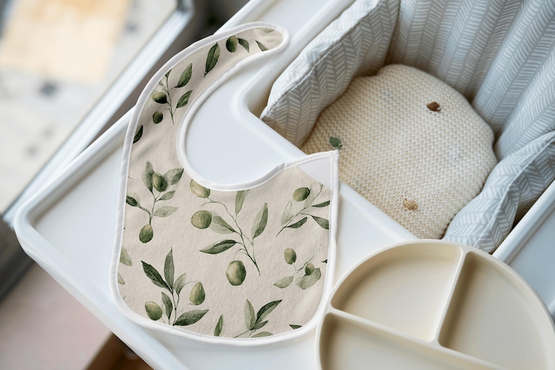 May include: A beige baby bib with a watercolor olive branch pattern, featuring green olives and leaves. The bib is on a white high chair tray, next to a divided plate and a cream-colored pillow.