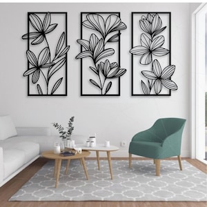 May include: Three black metal floral wall art panels. Each panel features a different stylized flower design within a rectangular frame. The artwork is displayed on a white wall above a living room setting with a sofa, chair, and coffee tables.