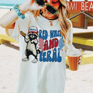 Op de afbeelding: Cr&egrave;mekleurig T-shirt met een afbeelding van een wasbeer met een "Merica"-hoed en een hotdog. Het shirt heeft de tekst "Red, White, and Feral" in rood, wit en blauw. De wasbeer draagt Crocs.