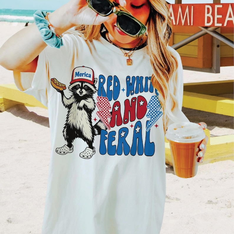 Op de afbeelding: Cr&egrave;mekleurig T-shirt met een afbeelding van een wasbeer met een "Merica"-hoed en een hotdog. Het shirt heeft de tekst "Red, White, and Feral" in rood, wit en blauw. De wasbeer draagt Crocs.